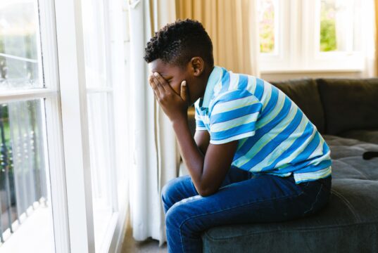 Sad african american boy covering his face and sitting at window in living room